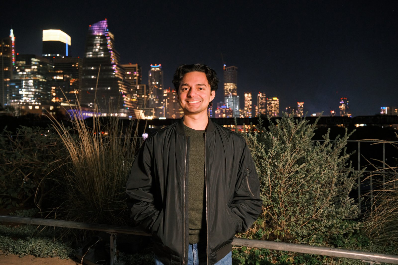 Photo of Michael Rolon, online alias "midilor", standing with the city of Austin skyline behind him. Wearing a black bomber jacket, jeans, and a green shirt, it's clear that he has an eye for aesthetic.